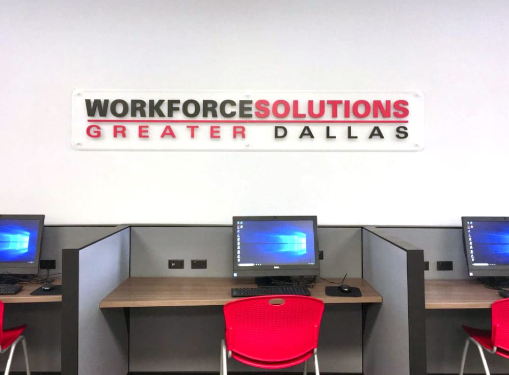 workforce-solutions A row of computer workstations with red chairs is set up in a modern office environment. Above the workstations, a sign on the wall reads "Workforce Solutions Greater Dallas" in bold letters. Each workstation has a computer monitor displaying the Windows desktop screen, indicating that the space is designed for public use, likely for job searching or workforce development activities. The setup is clean and organized, reflecting a professional and welcoming atmosphere.